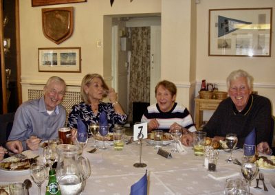A group of East Coast Branch Moody Owners sitting at a lunch table in front of a wall. Hanging on the wall are various pictures and plaques relating to Medway Yacht Club.